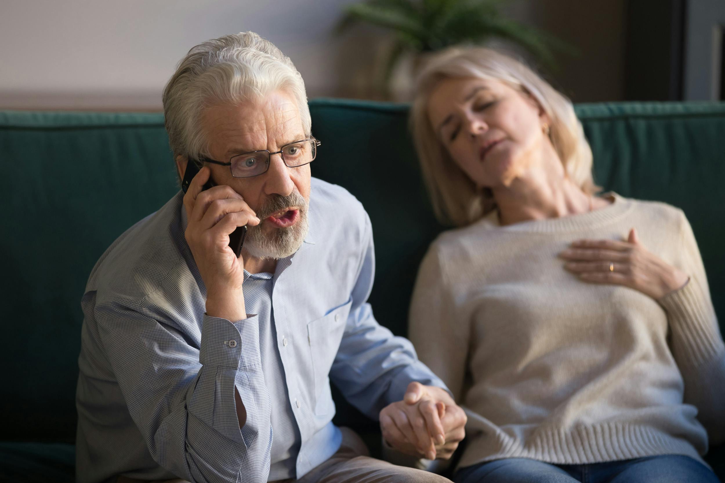 Worried elderly man talking on the phone while holding the hand of a sleeping or unwell-looking elderly woman.