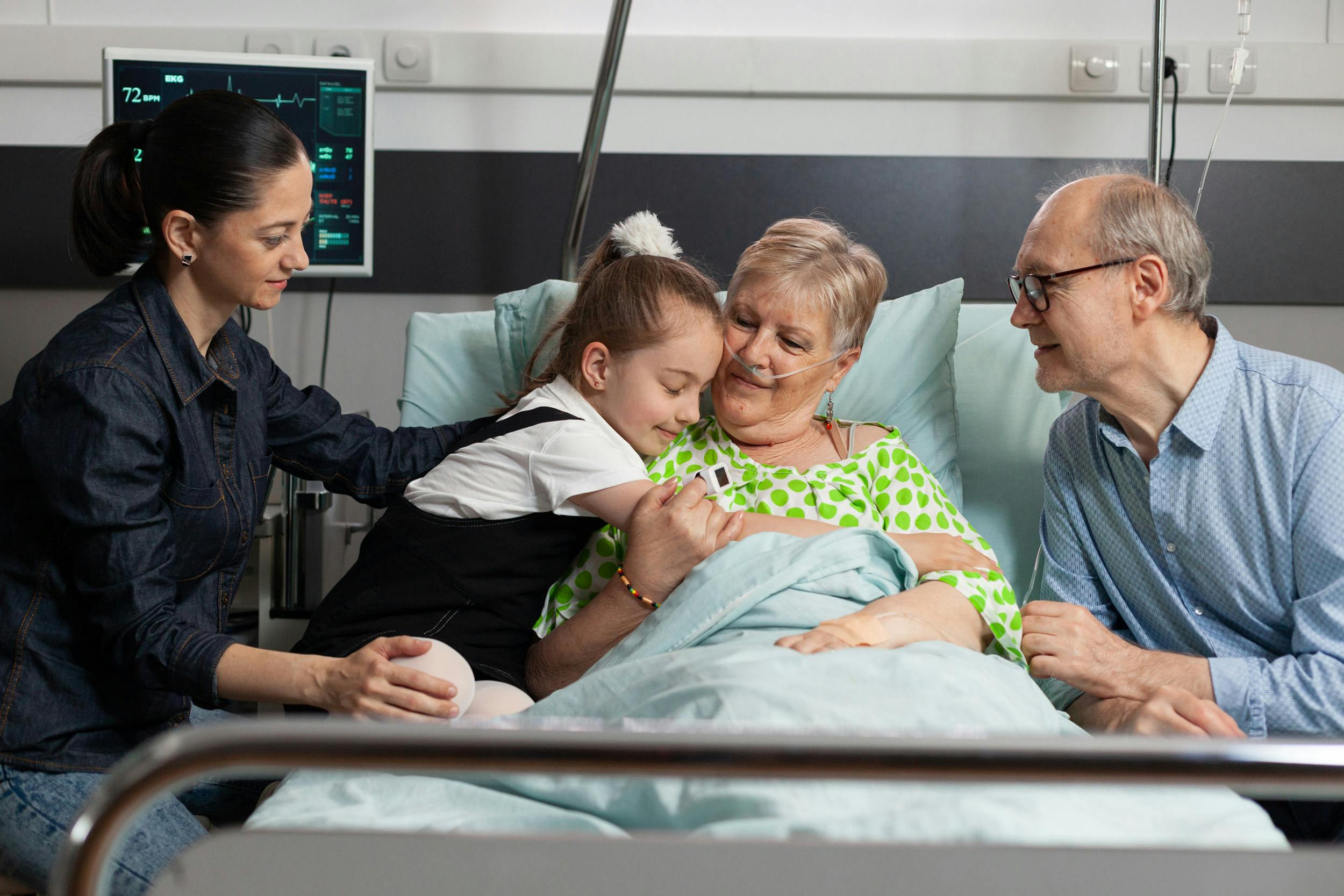 Ältere Frau mit Sauerstoffschlauch im Krankenhausbett hält ihre Enkelin, während ein älterer Mann liebevoll zusieht.