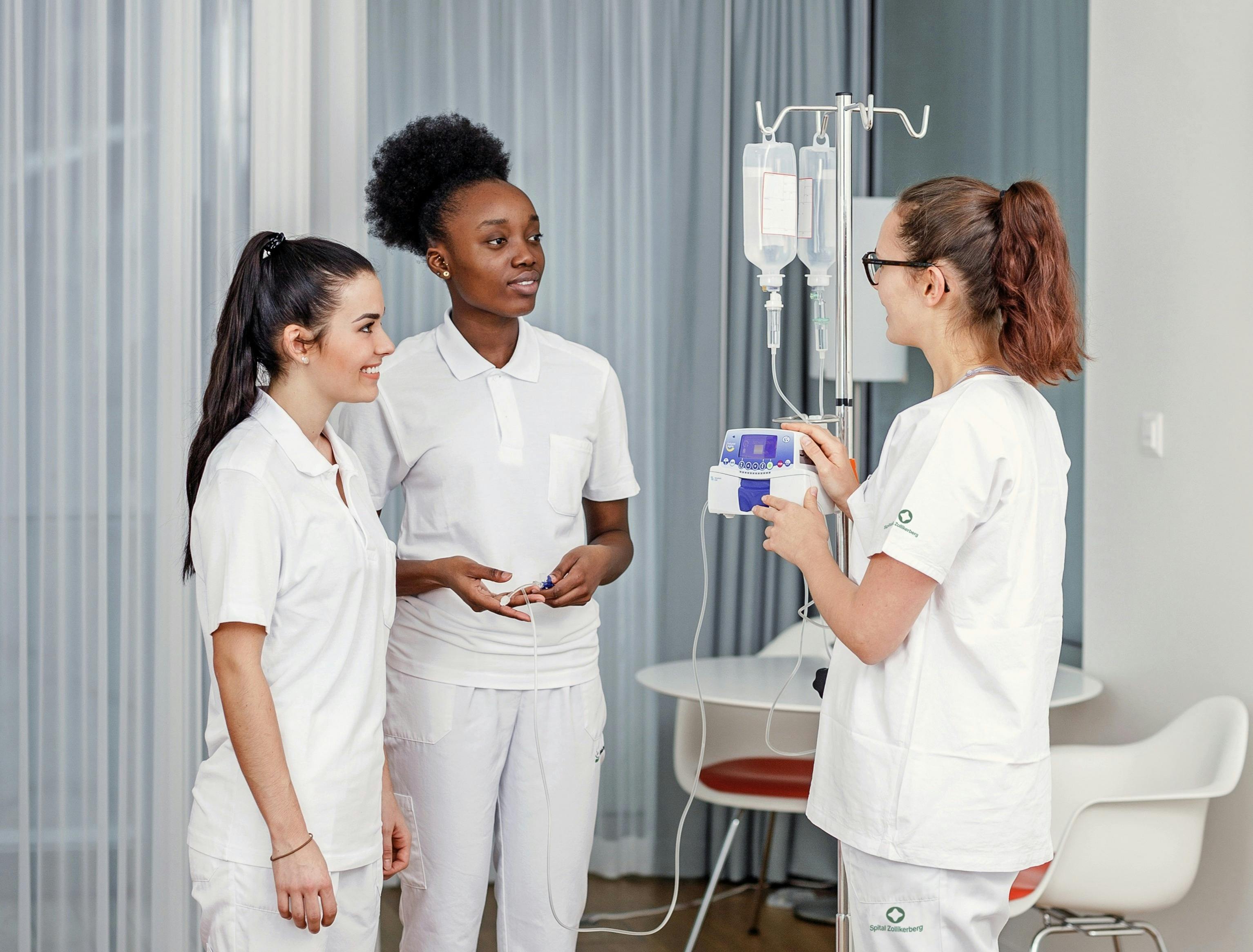 Drei Krankenpflegerinnen in weisser Berufskleidung unterhalten sich in einem Spitalzimmer.