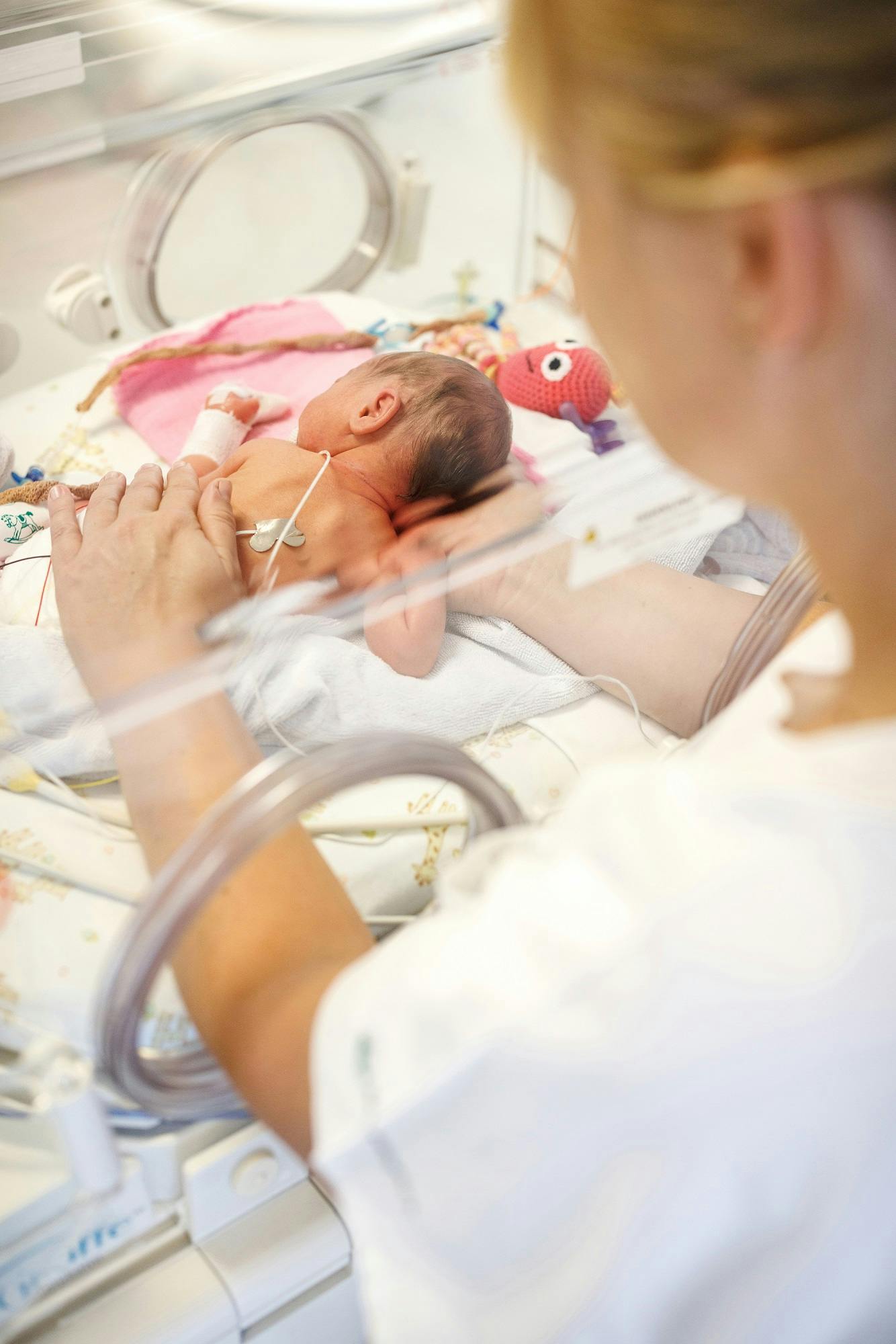 Frühgeborenes Baby in einem Inkubator mit medizinischen Geräten, versorgt von einer Person in weißer Kleidung.