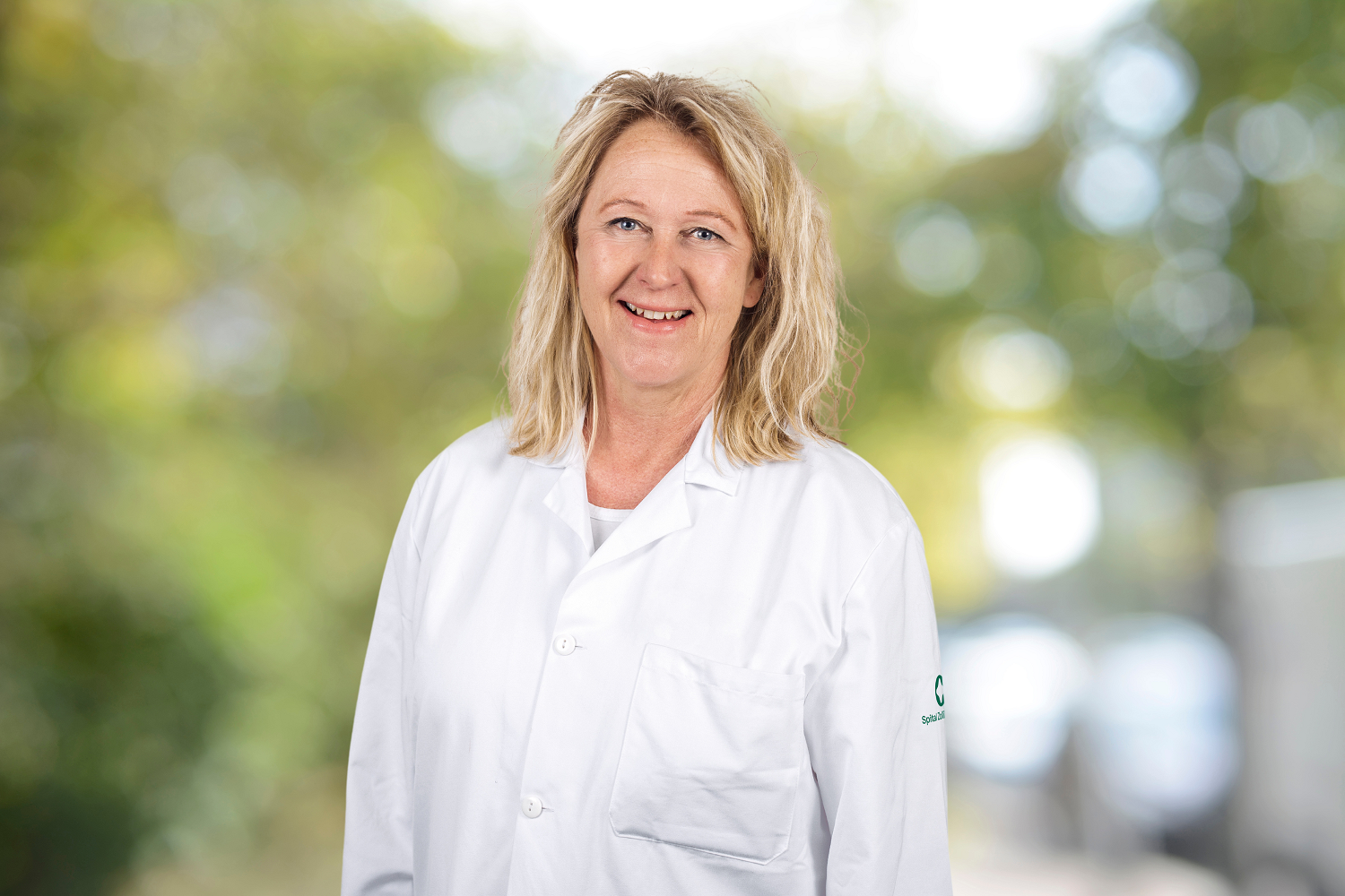 Woman in a white doctor's coat smiles in front of a blurred natural background.