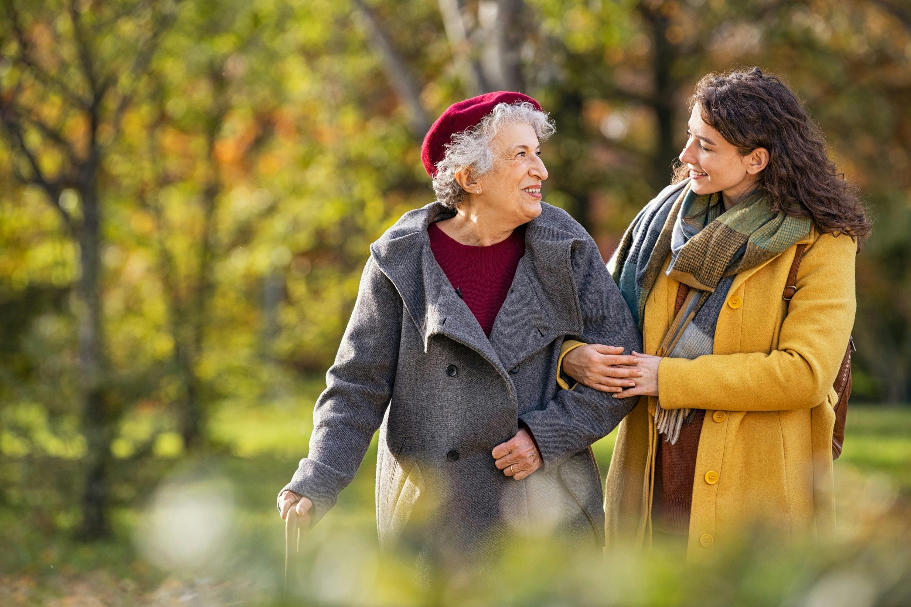 An older woman in a red cap and winter coat is walking through the forest in autumn next to a younger woman in a yellow coat. The two women smile at each other. 