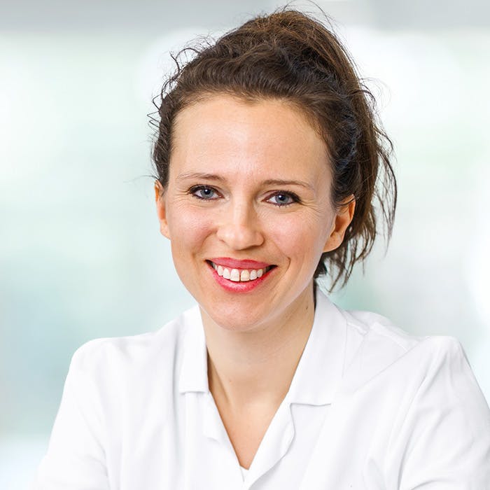 Smiling woman with a white coat in front of a blurred background.