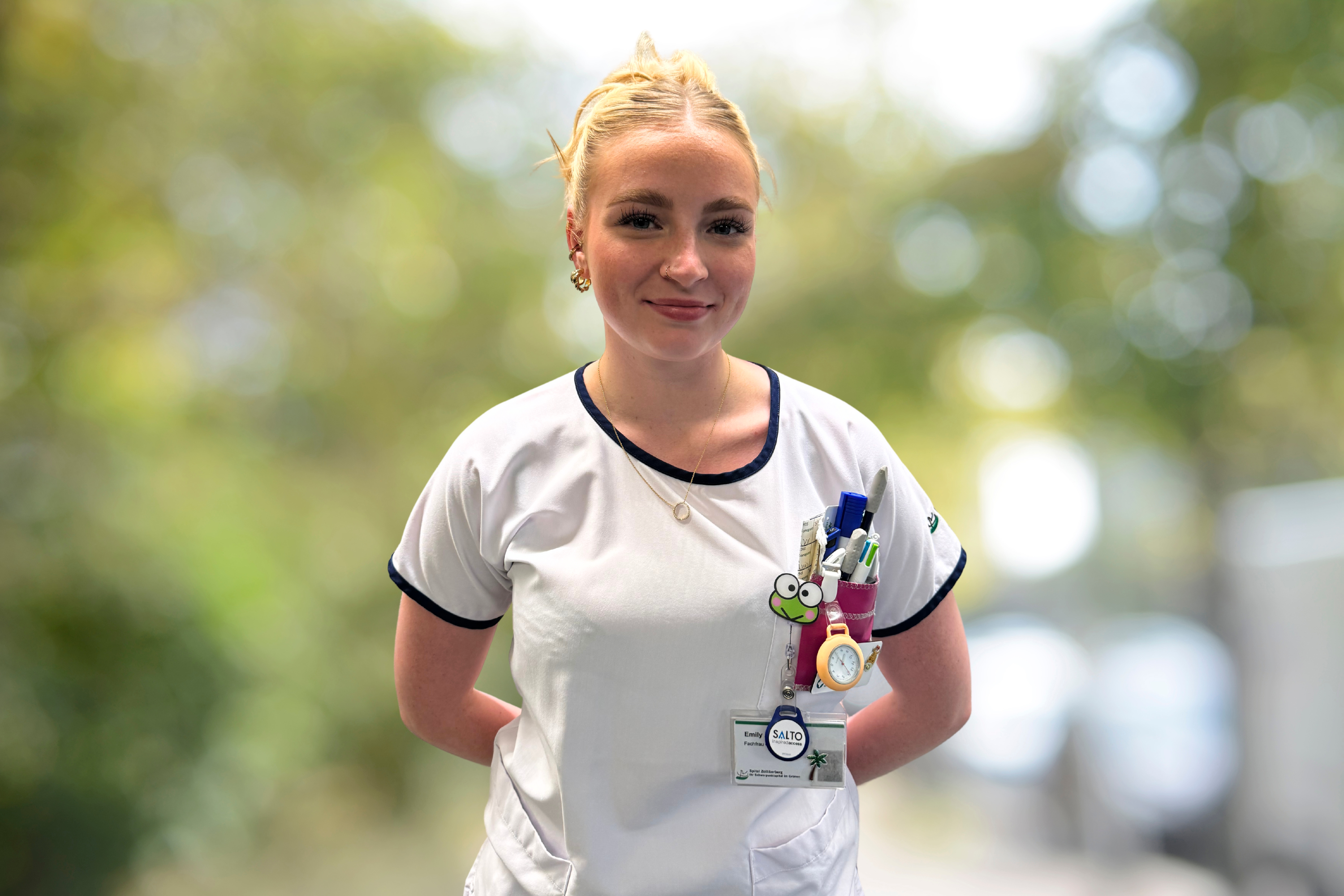 Smiling nurse with name tag and colourful pens in her pocket in front of a blurred background.