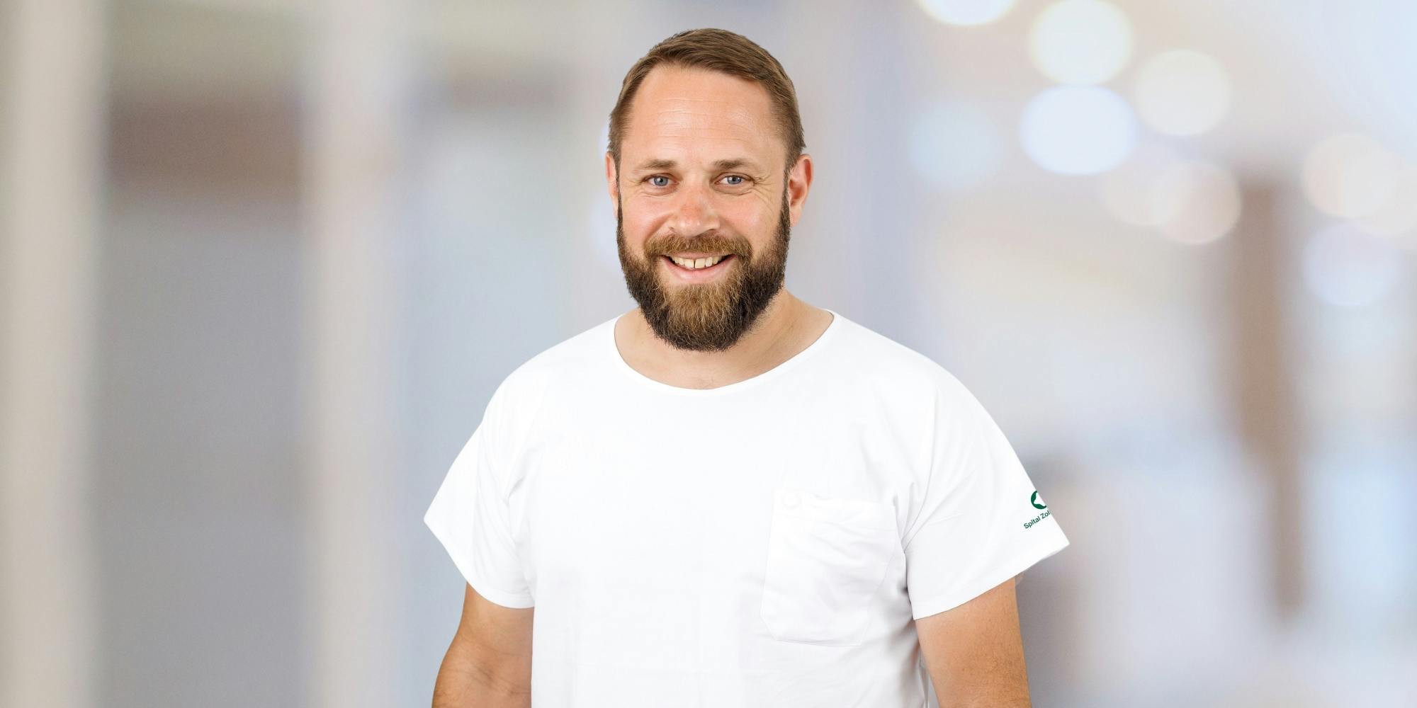 Smiling man with a beard in a white T-shirt against a blurred background.