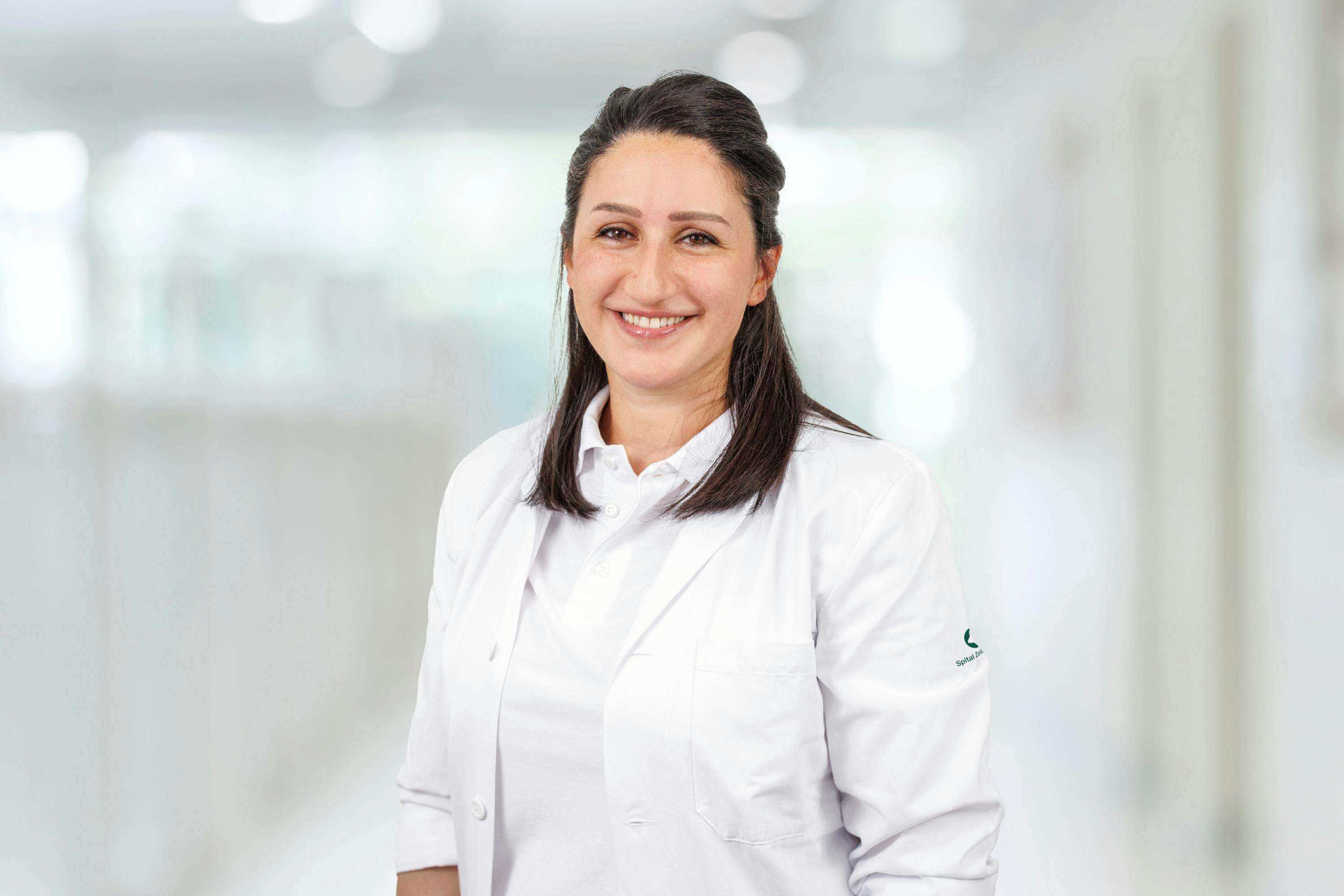 Lächelnde Frau im weißen Arztkittel vor unscharfem Hintergrund.
