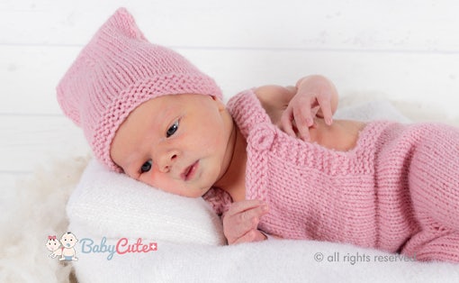 Neugeborenes mit rosa Mütze und Strampler auf weißer Unterlage.