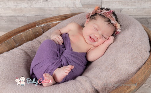 Newborn baby in purple wrap smiles in sleep with flower hairband.