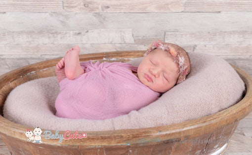 Newborn baby sleeping in a pink swaddle with headband, in a rustic wooden bowl.