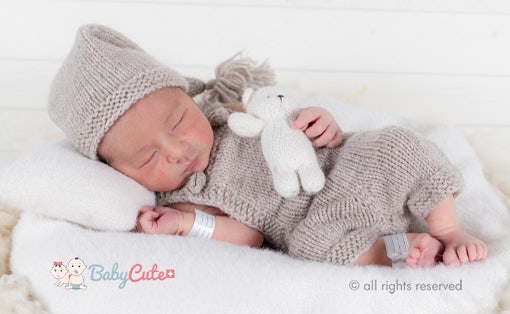 Schlafendes Neugeborenes mit Mütze und Strickanzug hält ein Stofftier.