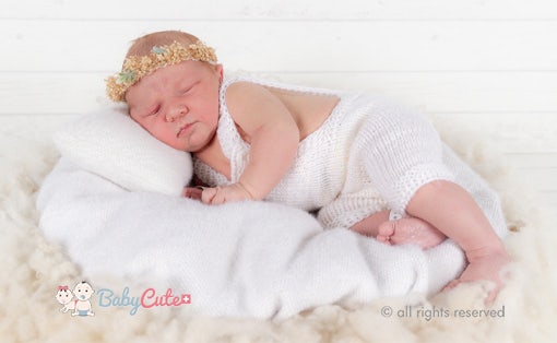 Neugeborenes schläft in weißem Strickoutfit mit Blumenhaarband.