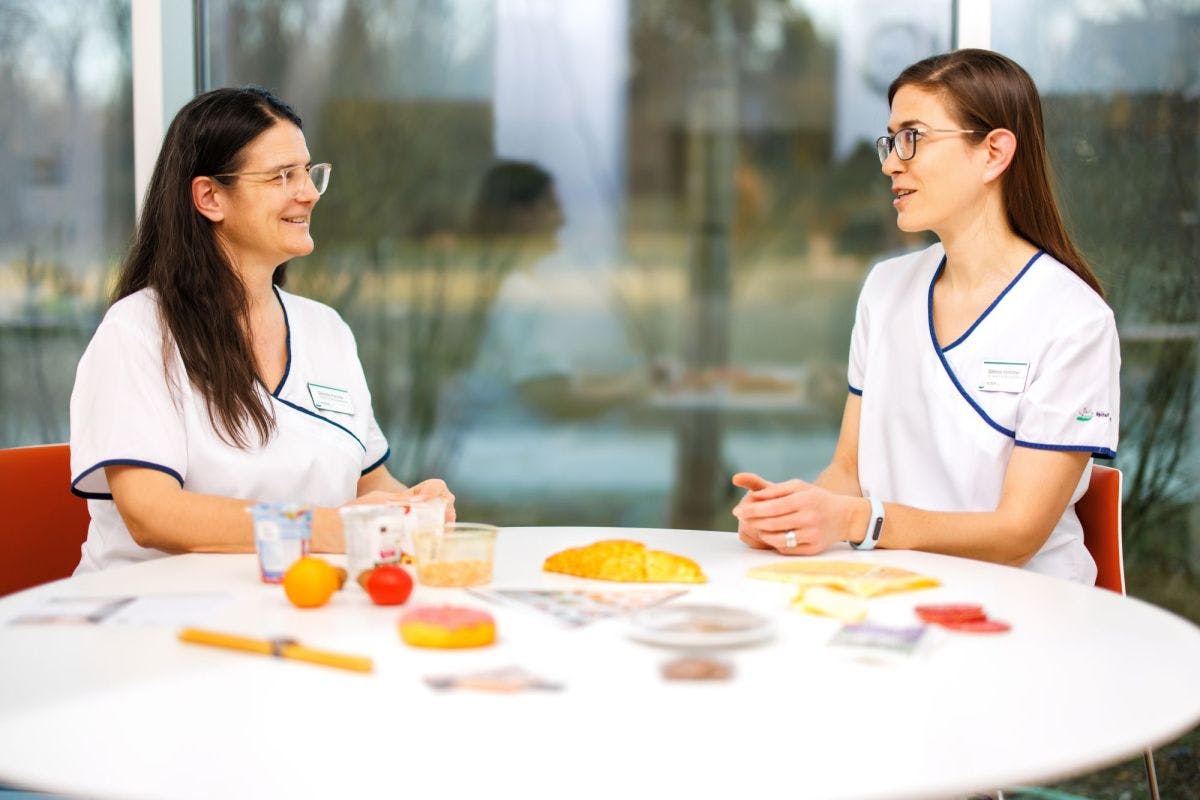 Two women in conversation during a coffee break at a conference with name badges and snacks on the table.