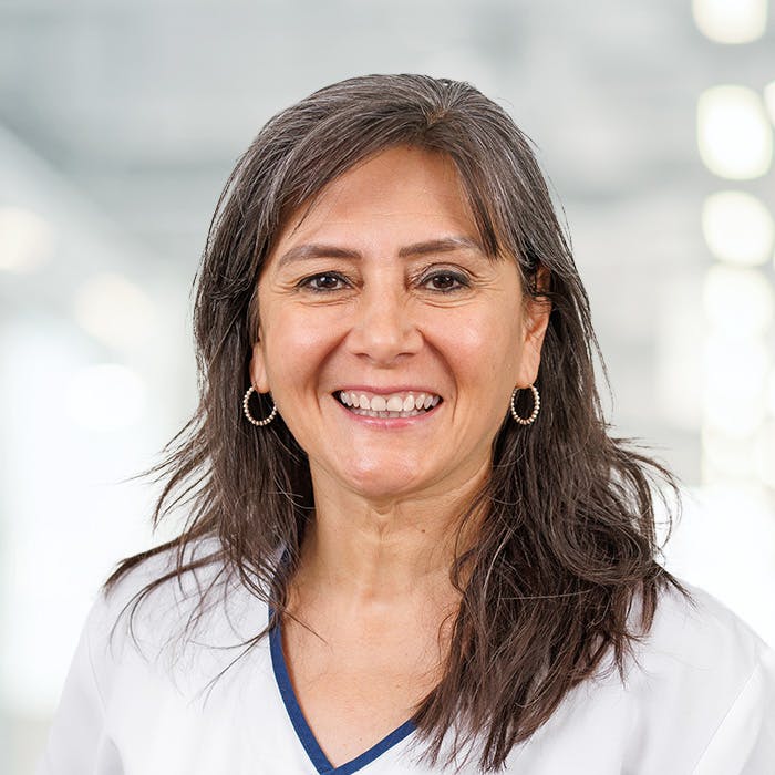 Smiling middle-aged woman with medical clothing and earrings against a blurred background.