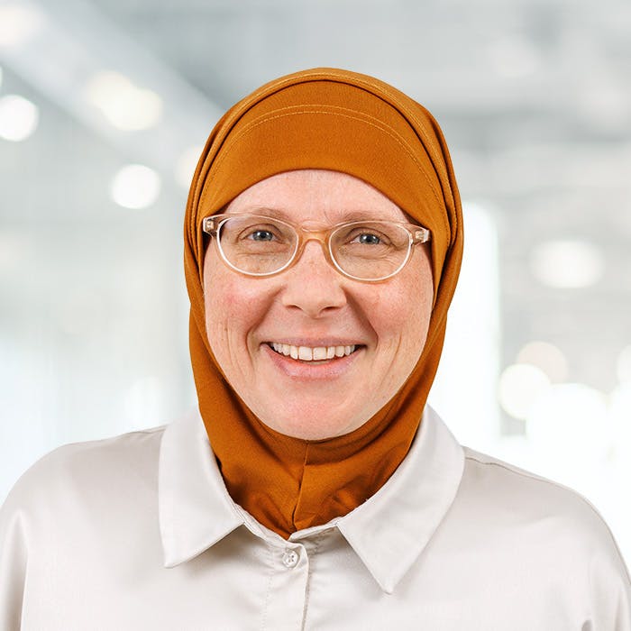 Woman with glasses and orange hijab smiling, against a blurred background.
