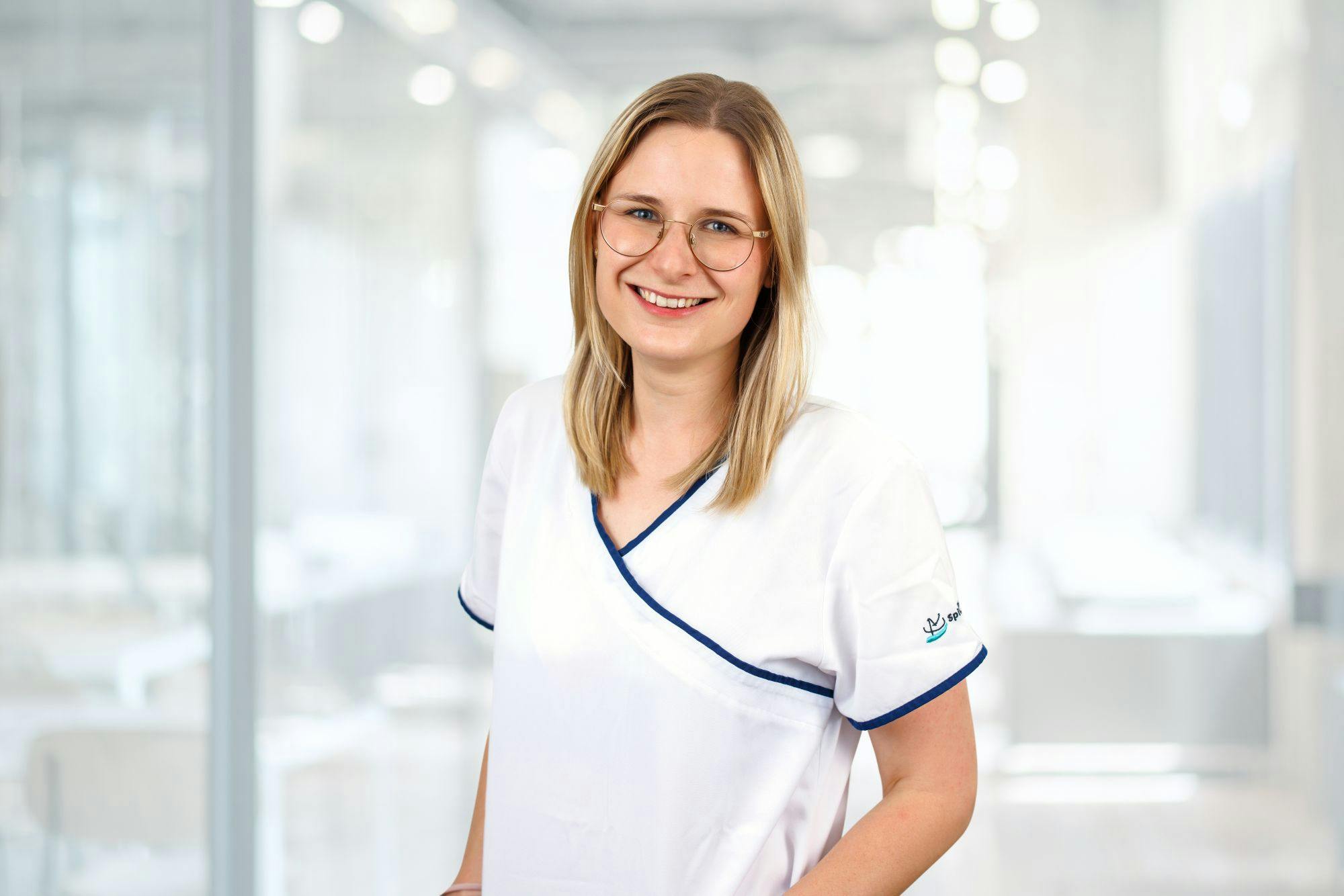 Junge lächelnde Krankenschwester im Berufskleid mit Brille vor unscharfem Klinik-Hintergrund.