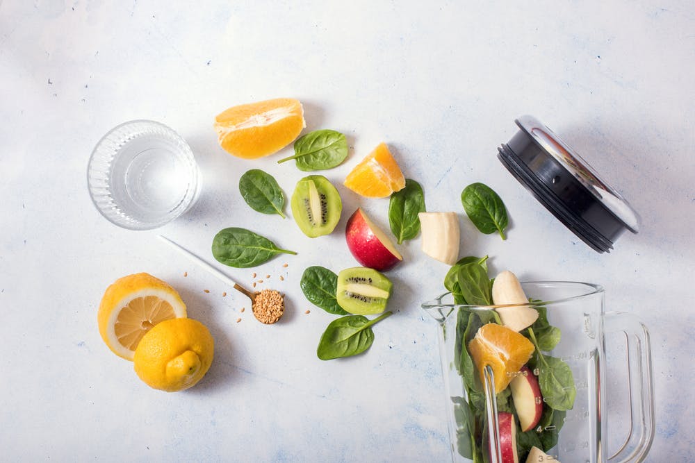 Fresh smoothie ingredients spread over a light-coloured surface, including lemon, kiwi, apple, spinach and a blender.