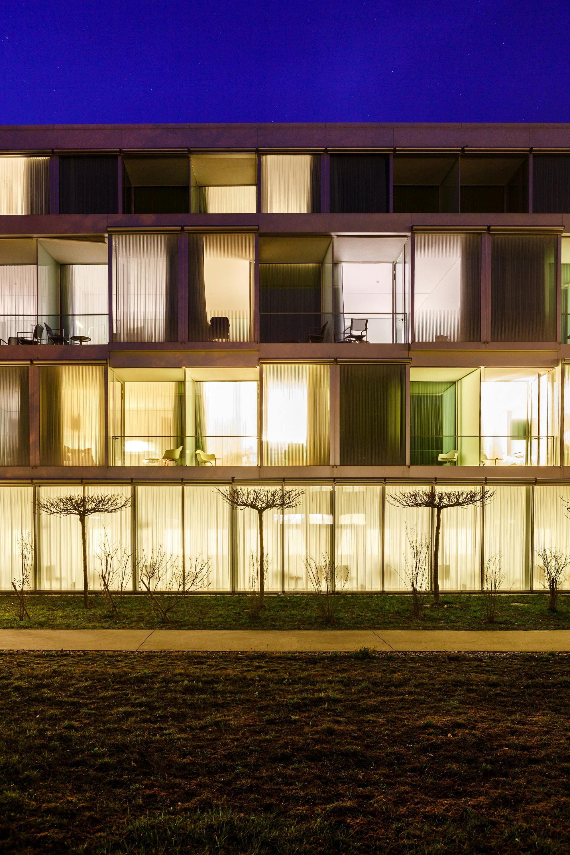 Beleuchtete Fassade eines modernen Wohngebäudes bei Nacht mit sichtbaren Innenräumen und Bäumen im Vordergrund.