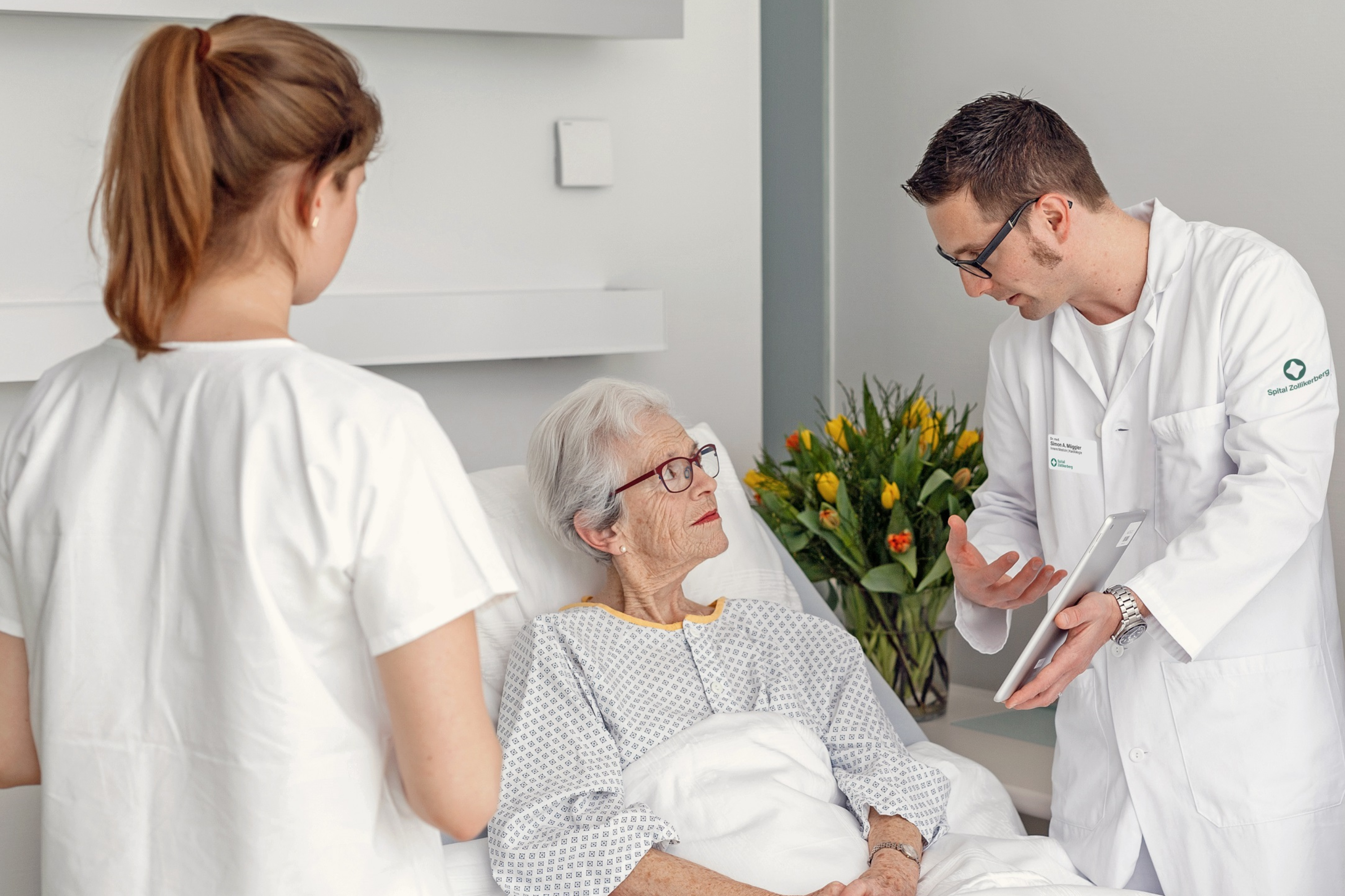 Ältere Patientin im Gespräch mit Arzt und Pflegerin im Krankenhauszimmer.