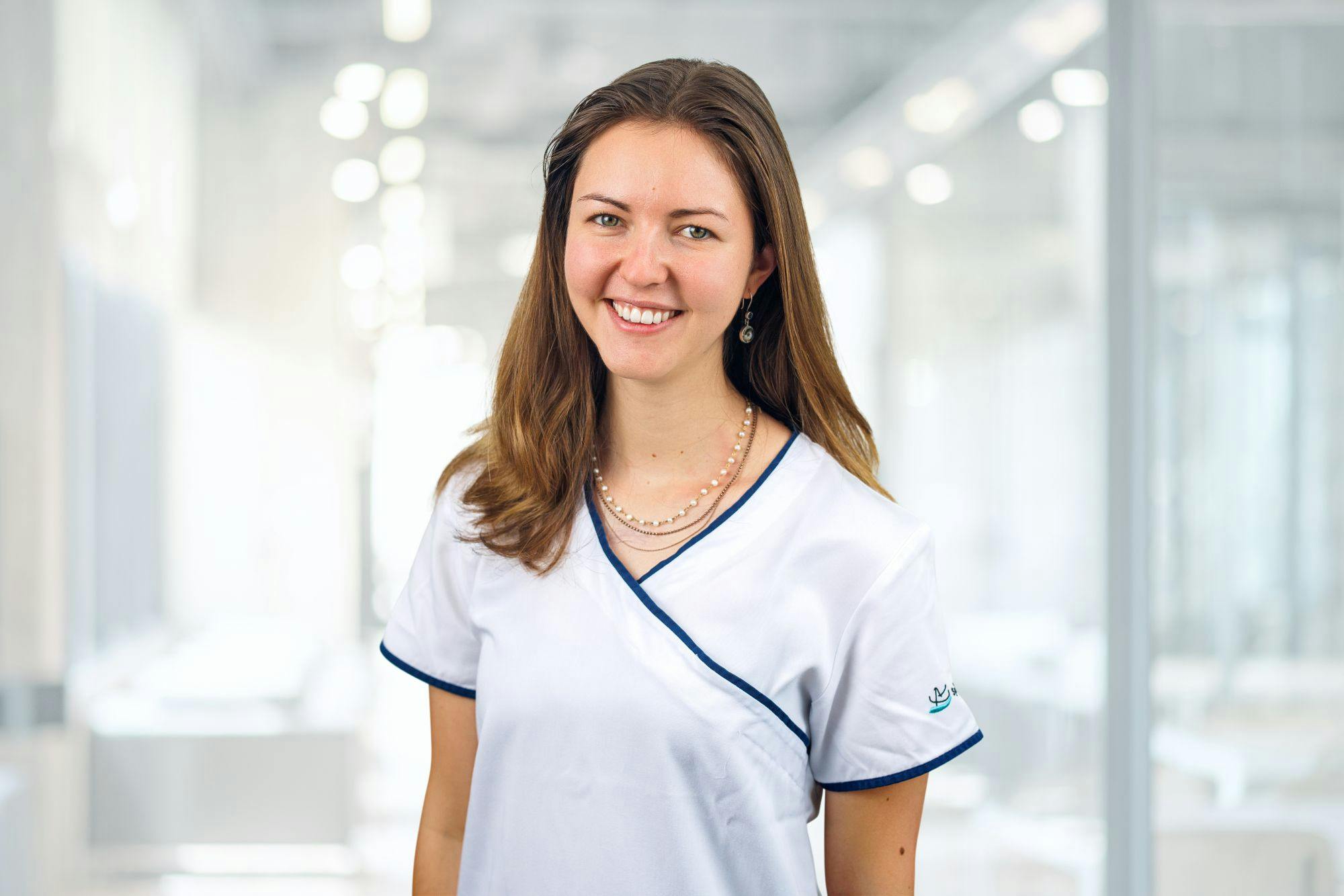 Lächelnde Frau in medizinischer Berufskleidung vor unscharfem Hintergrund.