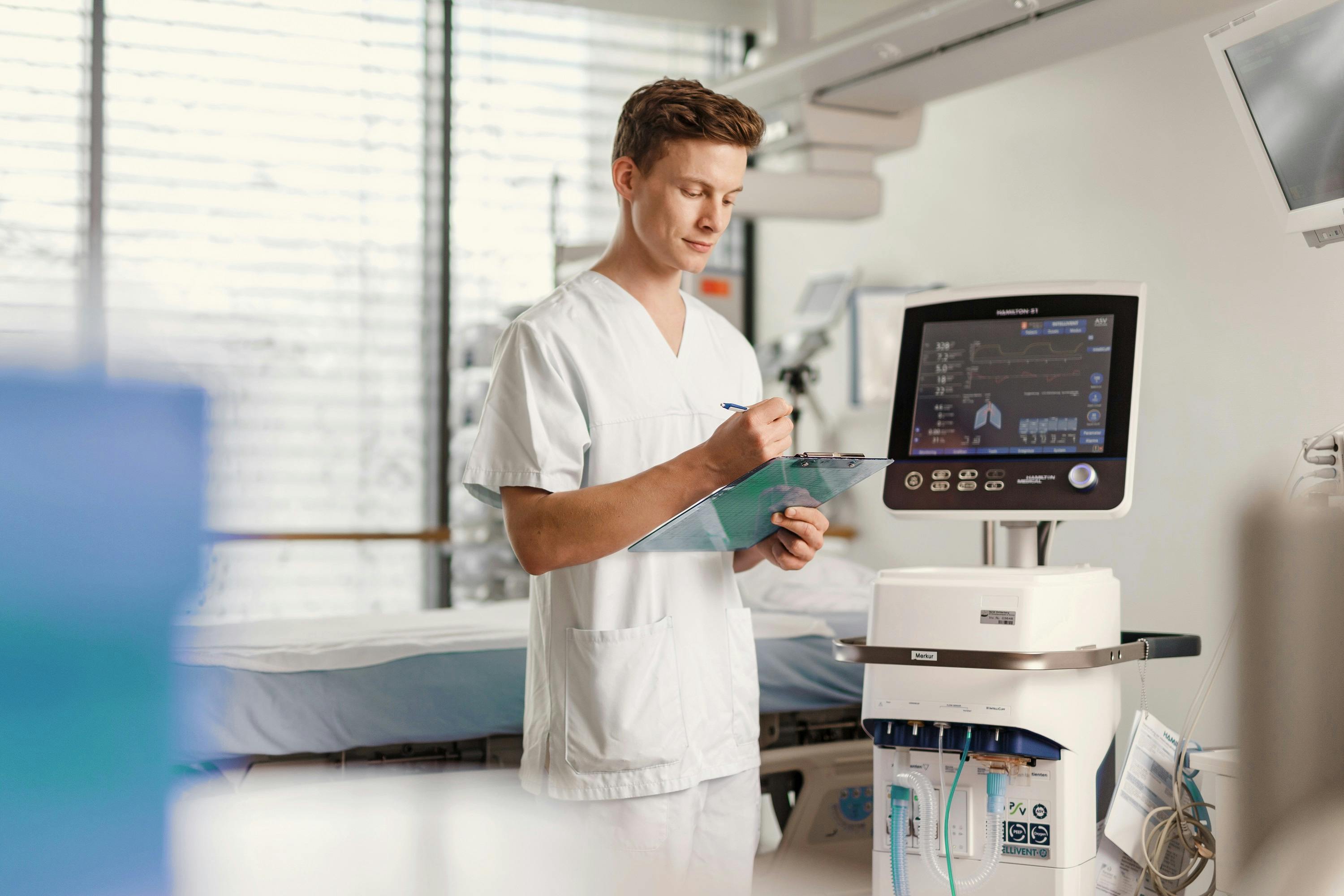 Medizinischer Fachangestellter in weißer Kleidung notiert Befunde im Krankenhauszimmer neben medizinischem Gerät.