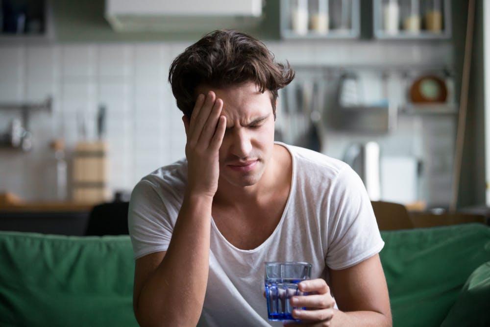 Junger Mann mit Kopfschmerzen hält Glas Wasser und stützt den Kopf ab.