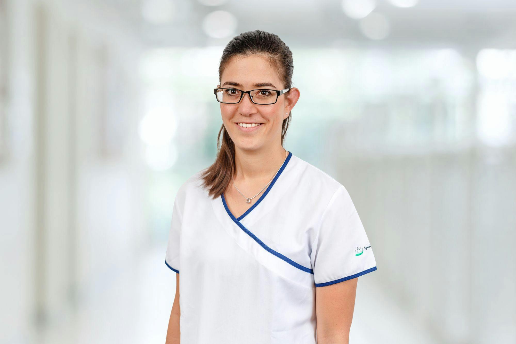 Lächelnde Medizinerin mit Brille und weißer Berufskleidung vor unscharfem Hintergrund.