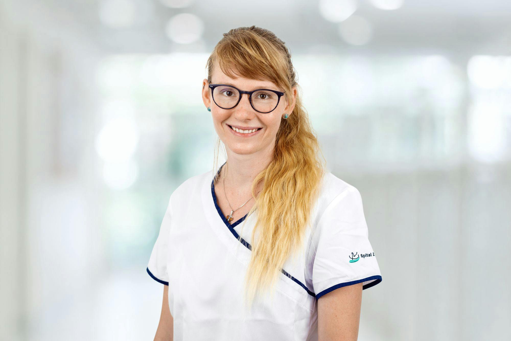 Lächelnde Frau in medizinischer Berufskleidung mit Brille und geflochtenem Haar.