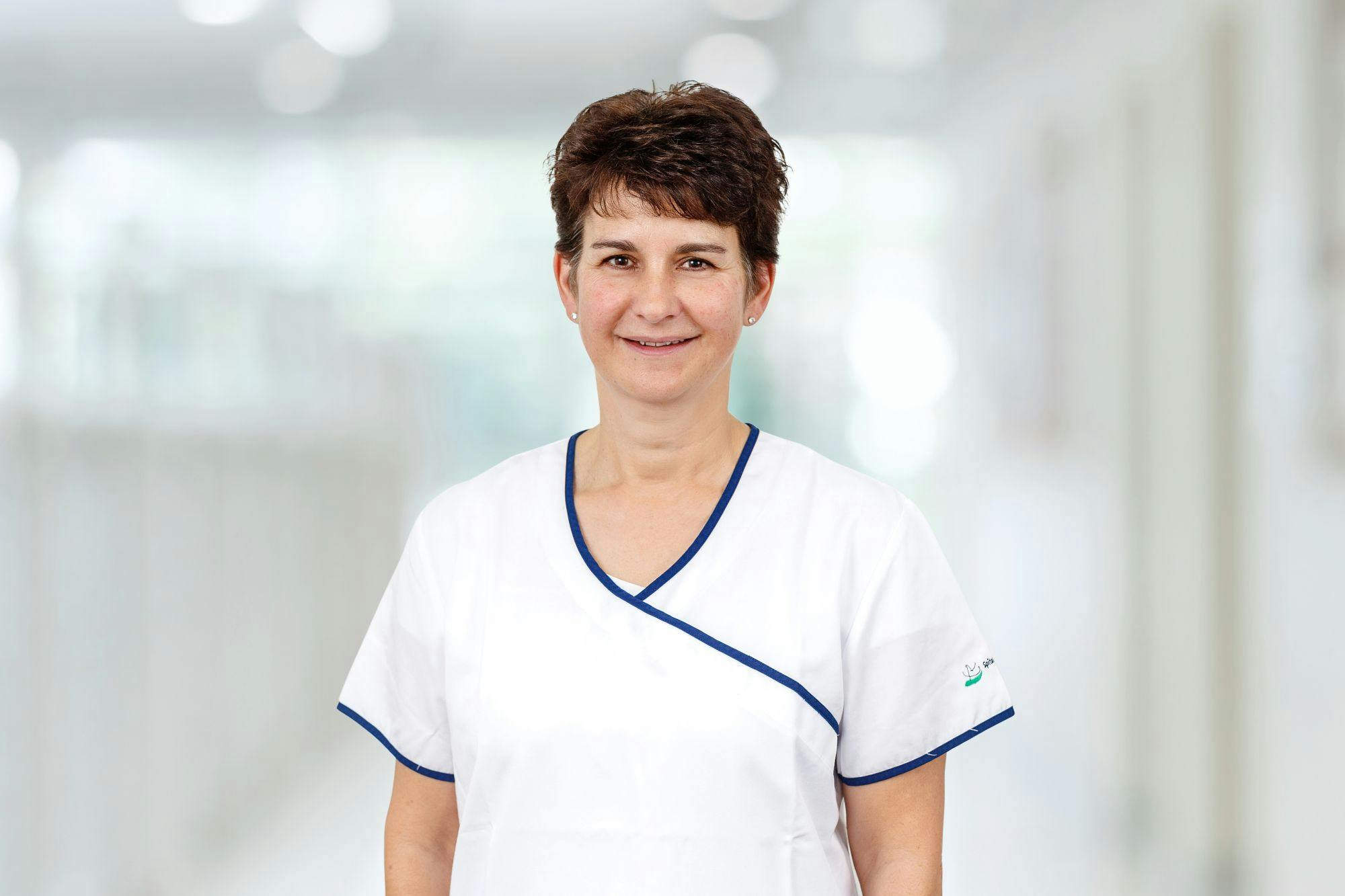 Smiling medical professional in white professional clothing with blue accents against a blurred background.