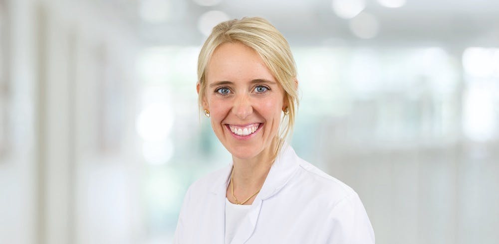 Lächelnde Frau in weißem Arztkittel vor unscharfem Hintergrund.