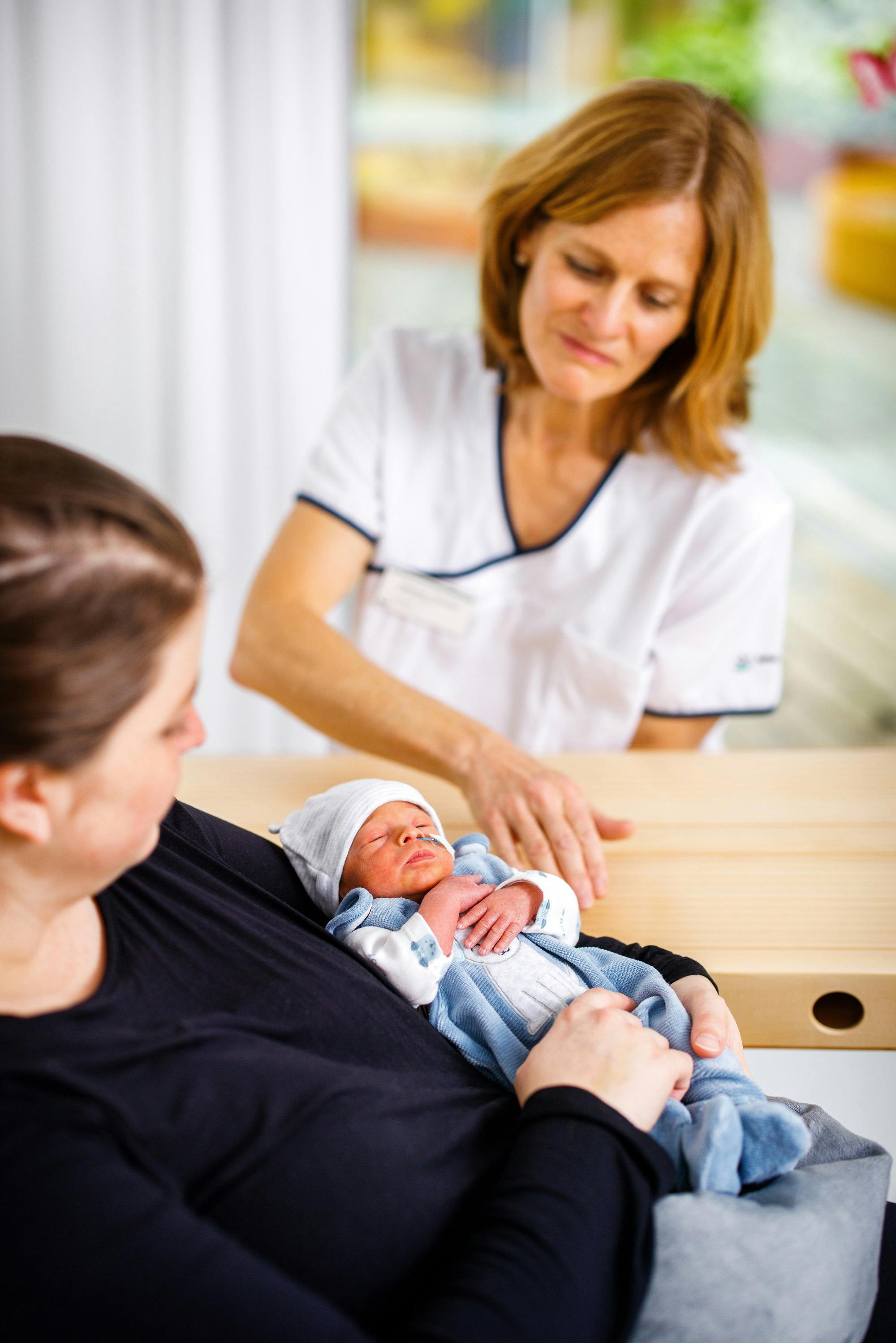 Neugeborenes auf dem Arm einer Frau mit Krankenschwester im Hintergrund.