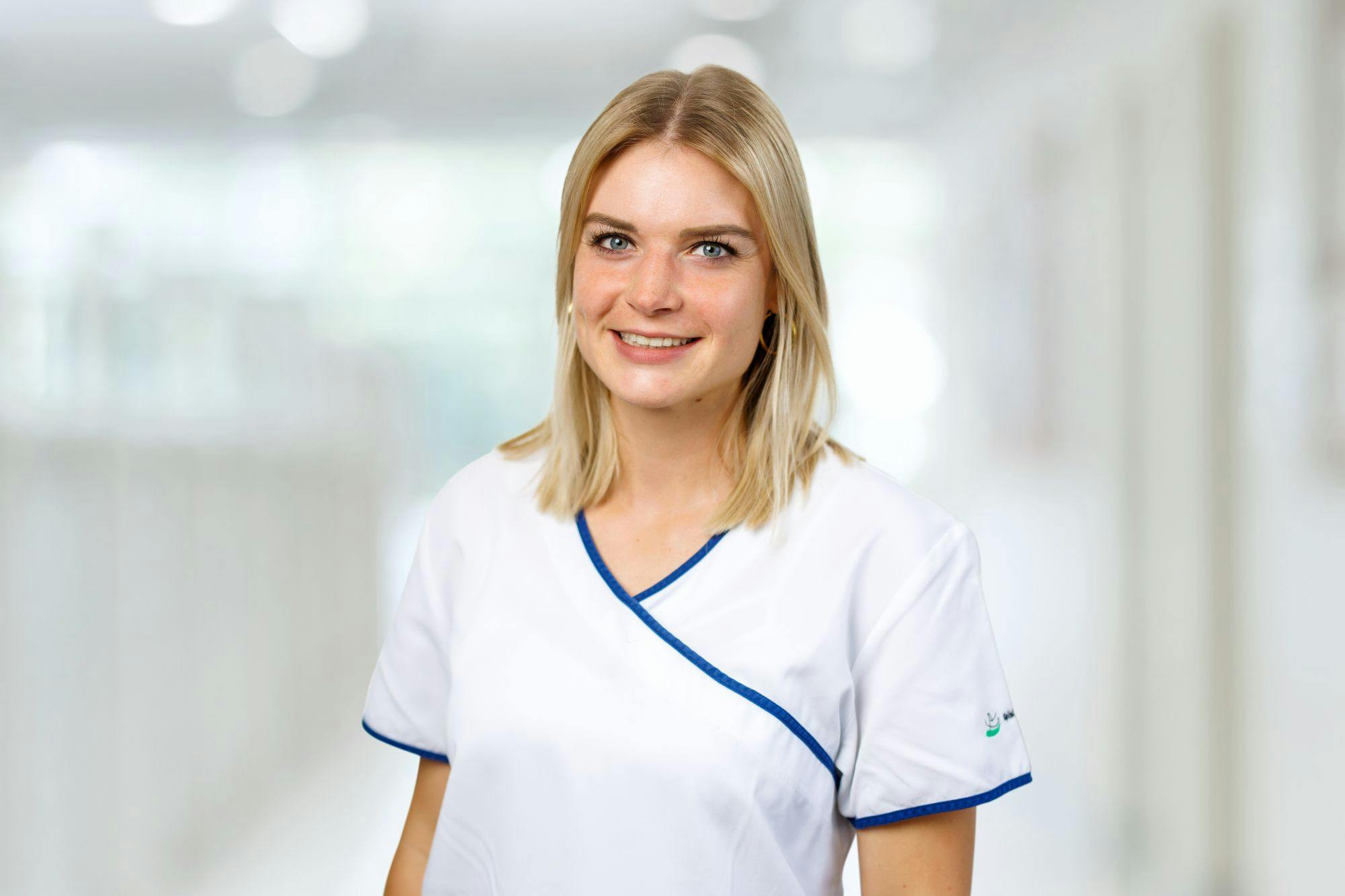 Junge lächelnde Frau in medizinischer Berufskleidung vor unscharfem Hintergrund.