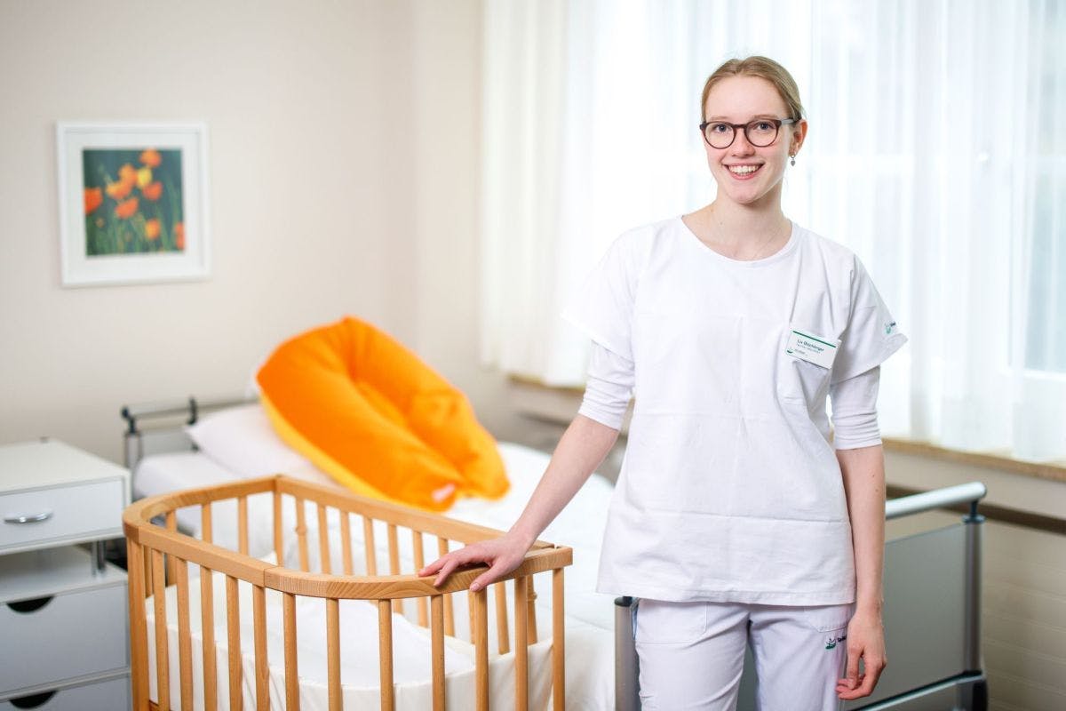 Lächelnde Pflegekraft im Krankenhauszimmer mit Kinderbett.