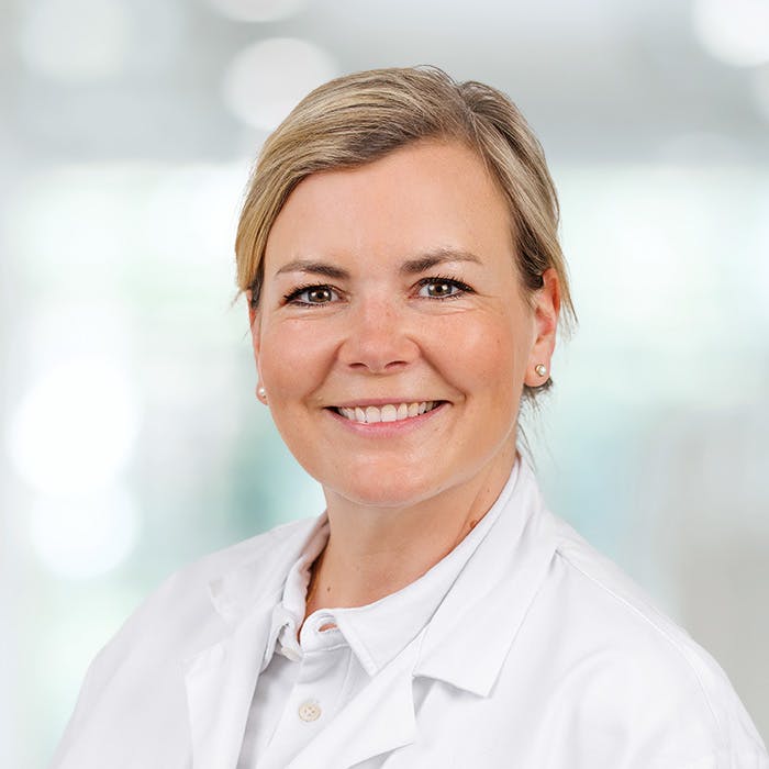 Smiling woman in a white coat, presumably a doctor, against a blurred background.