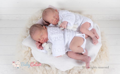 Zwei schlafende Neugeborene in weißen Strickoutfits, die auf einem weichen Kissen liegen.