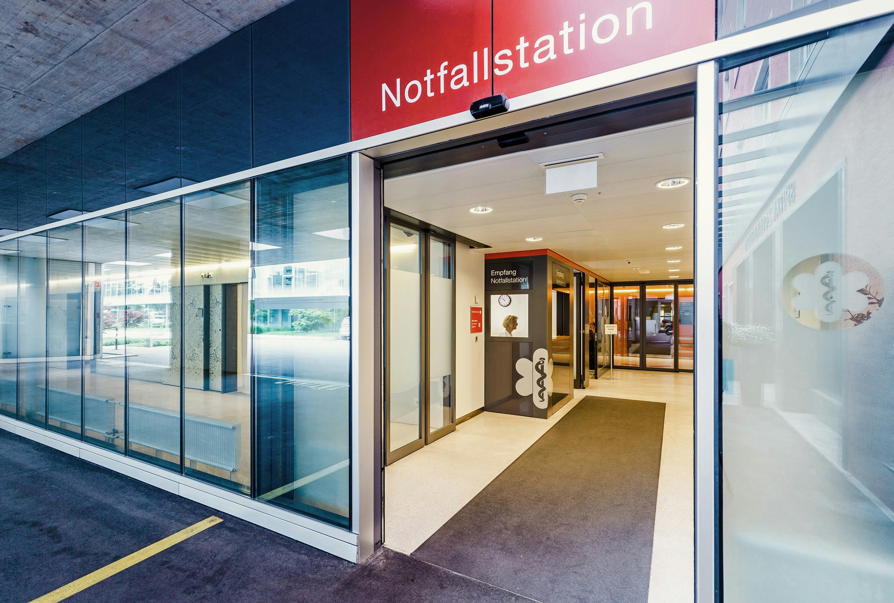 Eingang zur Notfallstation eines Krankenhauses mit geöffneten Glastüren und rotem Schild.