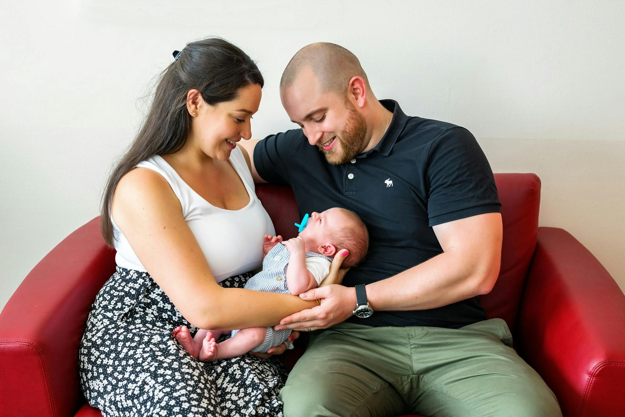 Glückliche junge Familie mit Neugeborenem auf rotem Sofa.